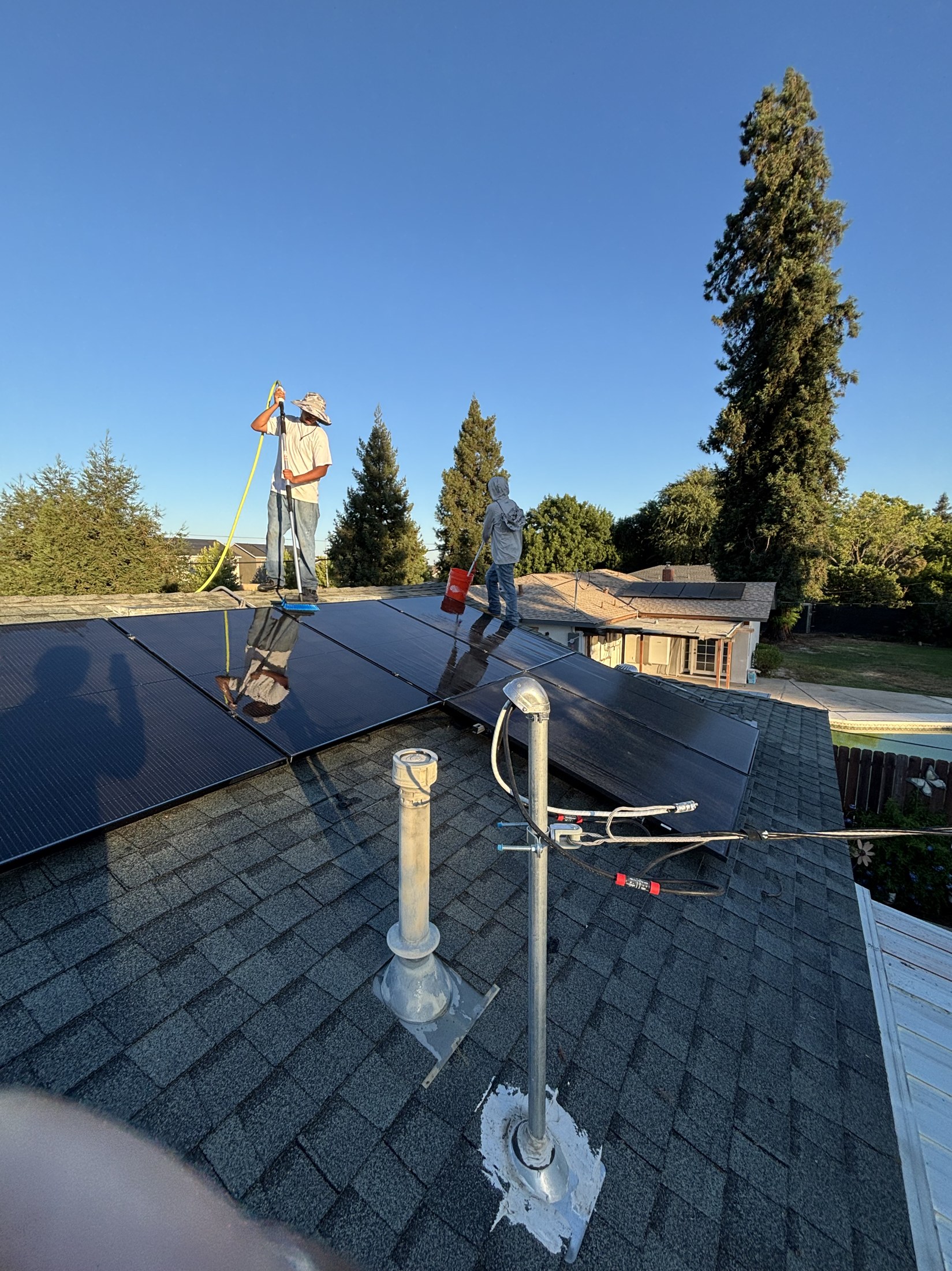 Technician cleaning rooftop solar panels with a water-fed brush pole under a clear blue sky.