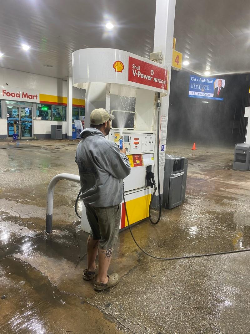 Technician pressure washing a Shell gas station fuel pump island at night to cut fuel and oil grime.